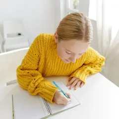 Dziewczynka w żółtym swetrze pisze w zeszycie, ucząc się, co to jest bezokolicznik.