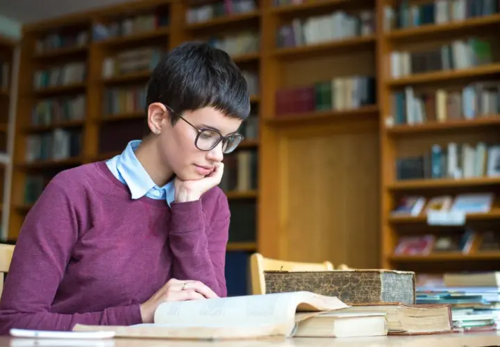 Młoda kobieta w okularach pochłonięta lekturą, szukająca odpowiedzi na pytanie: transcendencja co to? W otoczeniu bibliotecznych regałów.