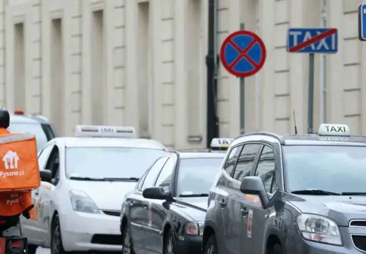 Kurier z pomarańczową torbą Pyszne.pl mija rząd samochodów taxi. Na ulicy widać znaki zakazu postoju.