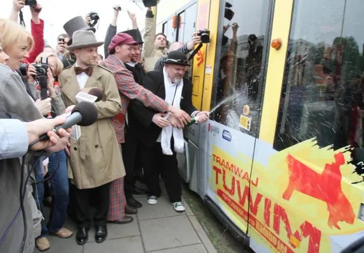 Mężczyzna w czarnym kapeluszu i białym szaliku otwiera szampana na tle tramwaju z napisem "Tramwaj Tuwim".
