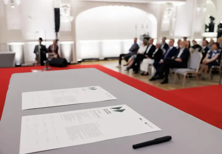 Na stole leżą dokumenty z napisem "SZLAK PRACY ORGANICZNEJ". W tle siedzą ludzie, czekając na rozpoczęcie wydarzenia, które symbolizuje pracę u podstaw.