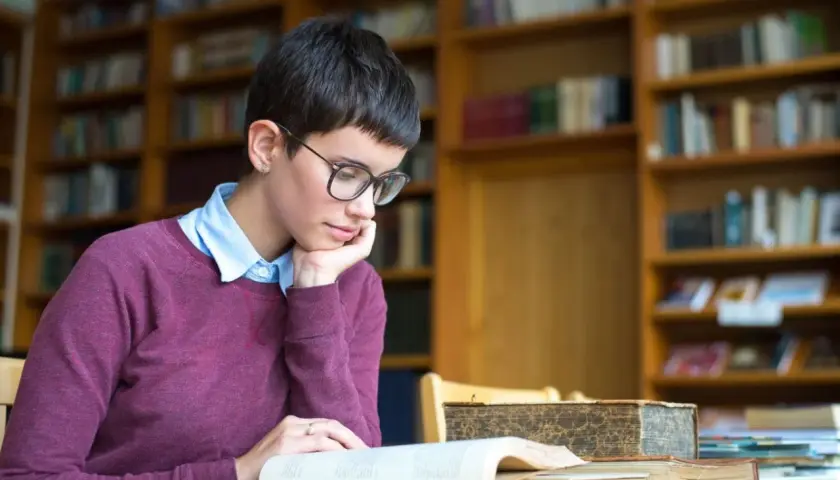 Młoda kobieta w okularach pochłonięta lekturą, szukająca odpowiedzi na pytanie: transcendencja co to? W otoczeniu bibliotecznych regałów.