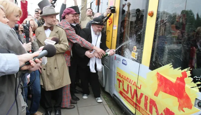 Mężczyzna w czarnym kapeluszu i białym szaliku otwiera szampana na tle tramwaju z napisem "Tramwaj Tuwim".