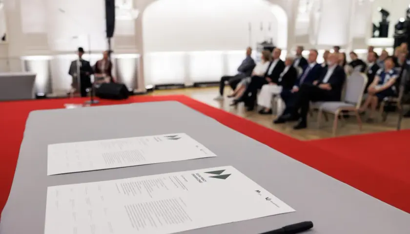 Na stole leżą dokumenty z napisem "SZLAK PRACY ORGANICZNEJ". W tle siedzą ludzie, czekając na rozpoczęcie wydarzenia, które symbolizuje pracę u podstaw.