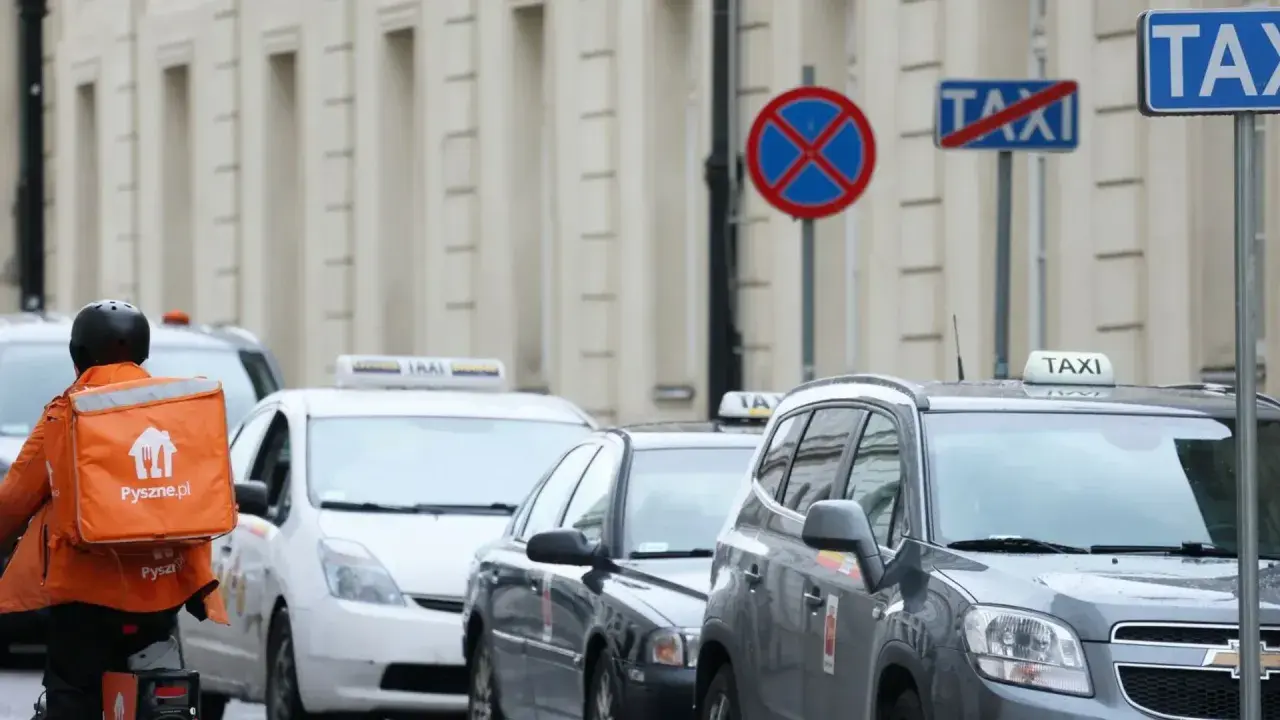 Kurier z pomarańczową torbą Pyszne.pl mija rząd samochodów taxi. Na ulicy widać znaki zakazu postoju.