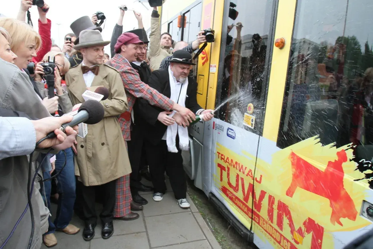 Mężczyzna w czarnym kapeluszu i białym szaliku otwiera szampana na tle tramwaju z napisem "Tramwaj Tuwim".