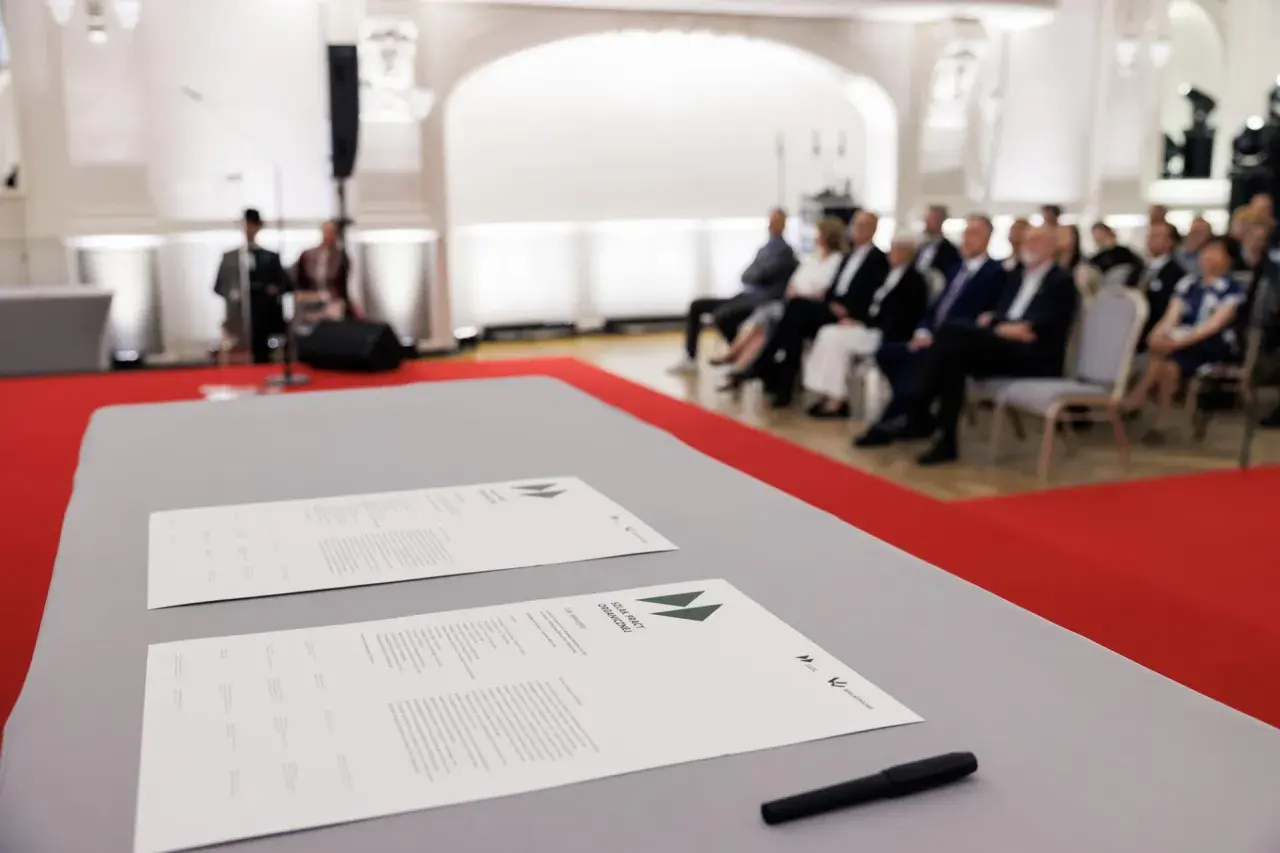 Na stole leżą dokumenty z napisem "SZLAK PRACY ORGANICZNEJ". W tle siedzą ludzie, czekając na rozpoczęcie wydarzenia, które symbolizuje pracę u podstaw.