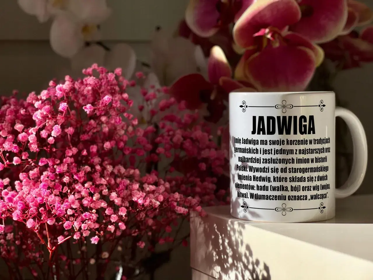 Biały kubek z napisem "Jadwiga" i opisem znaczenia imienia, na tle różowych kwiatów i storczyków.