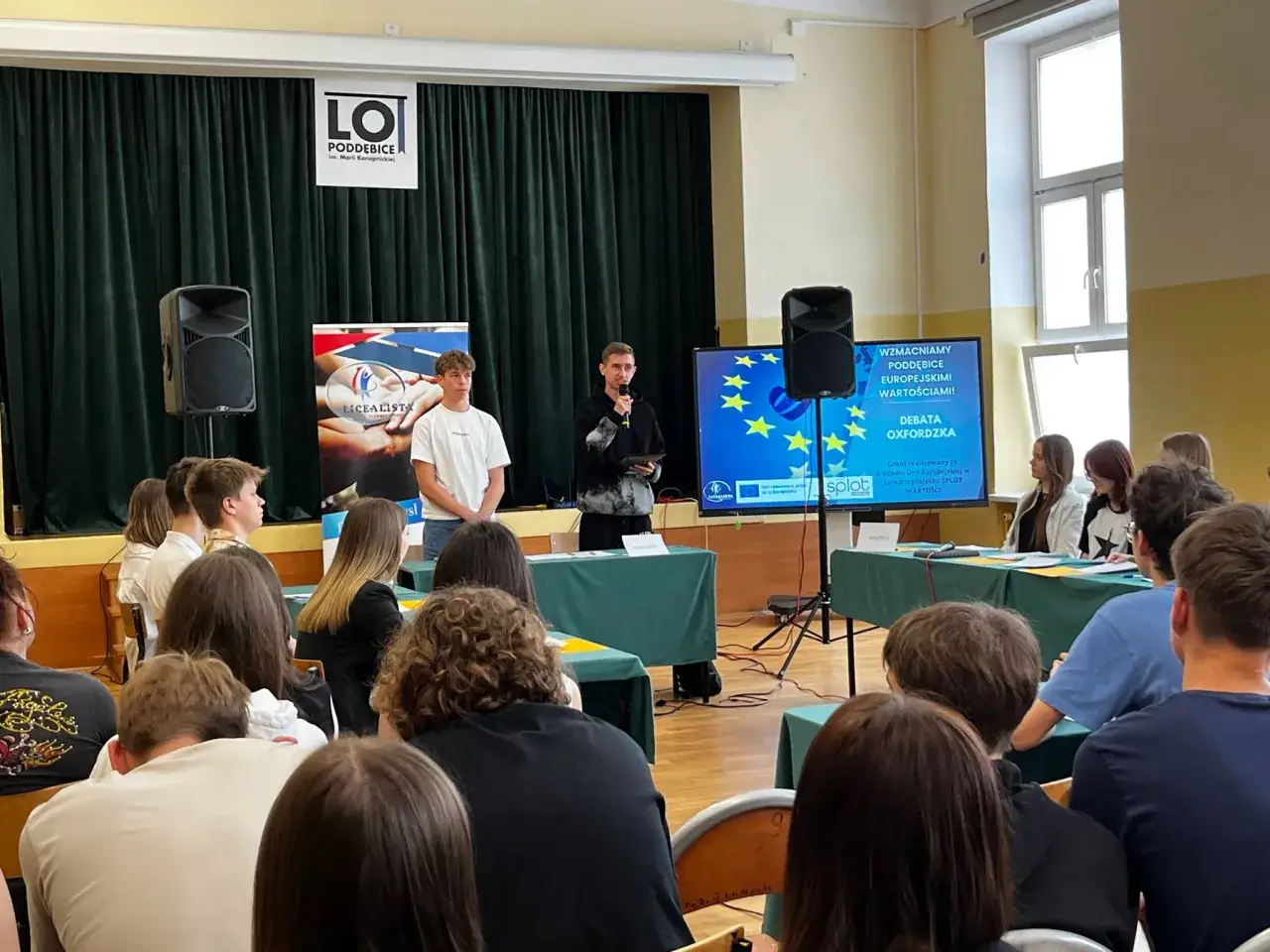 Młodzież w szkole podczas debaty oksfordzkiej. Jeden z uczniów przemawia, a na ekranie widać flagę UE. Kto to jest adwersarz? To ten, kto ma inne zdanie.