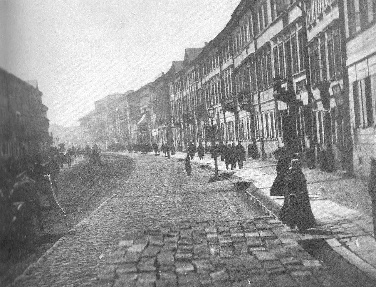 Ulica z kamienną nawierzchnią, szereg kamienic i ludzie w długich płaszczach. To jakby lalka czas akcji, zatrzymana w kadrze.