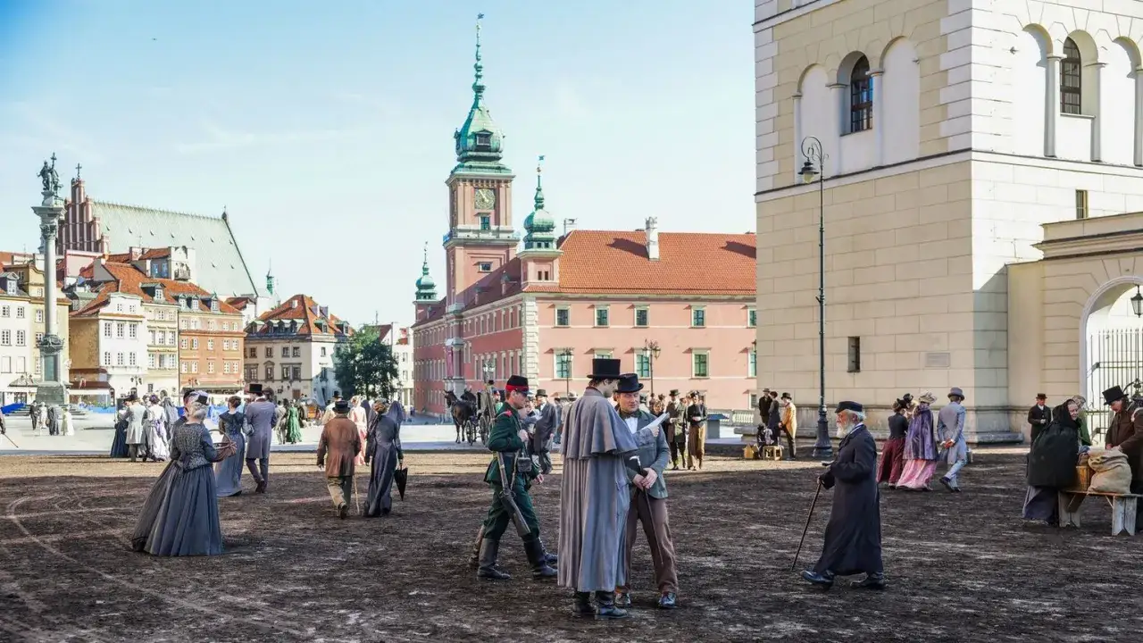 Scena z epoki, ludzie w strojach z XIX w. na placu. W tle zamek i kamienice. Praca u podstaw lalka, jak żywi.