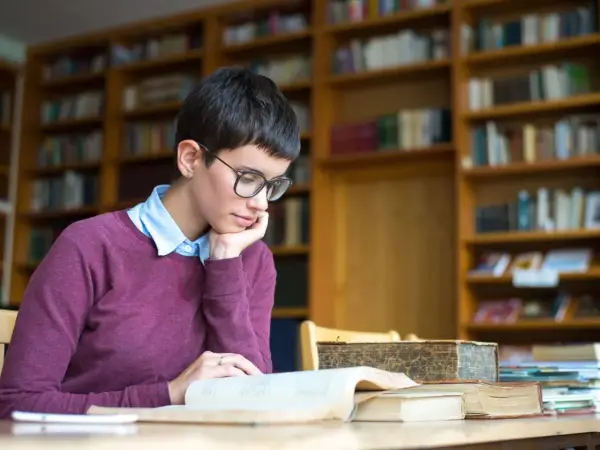Młoda kobieta w okularach pochłonięta lekturą, szukająca odpowiedzi na pytanie: transcendencja co to? W otoczeniu bibliotecznych regałów.
